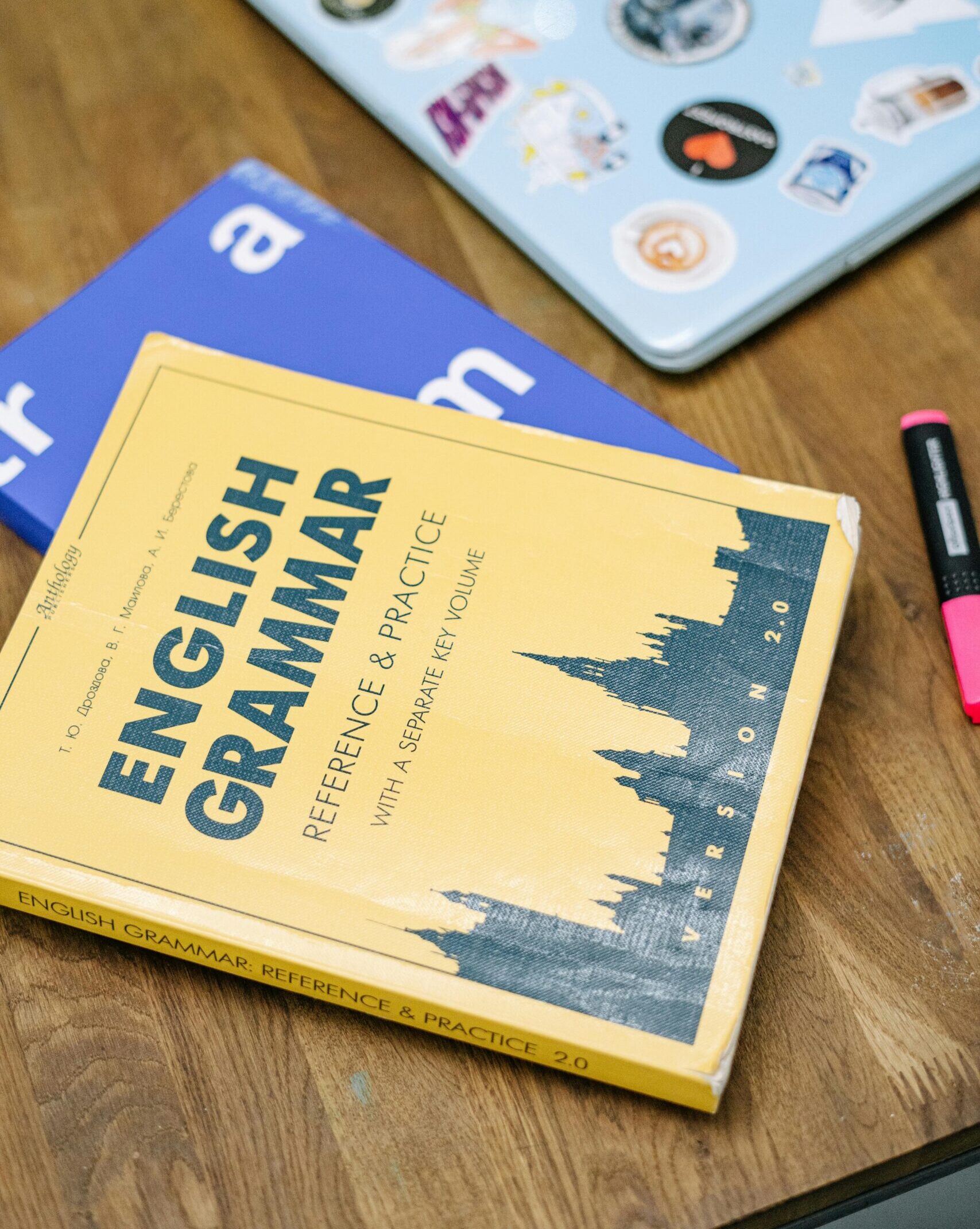 High-angle view of an English grammar book and a laptop with stickers on a wooden desk.